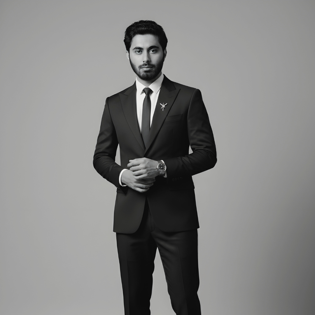 A man in a black suit posing with a thoughtful expression, his hand resting on his chin, against a soft grey studio background.